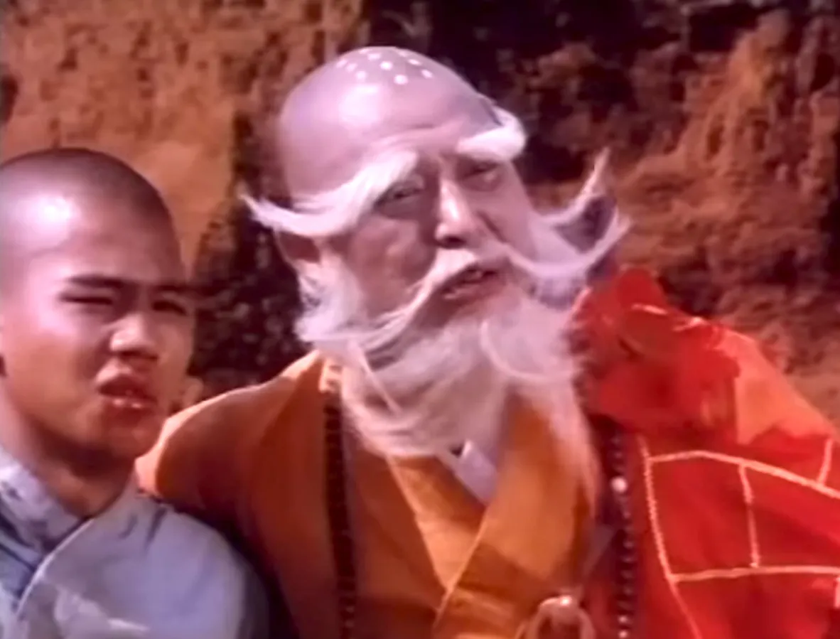 abbot-it-is-fated-show-him-mercy-let-him-go-in-peace-shaolin-vs-lama Image of Grandmaster and Shao Si Yer in a barren land with mountainous terrain and reddish sand. Grandmaster and Shao Si Yer are stand next to each other while looking toward the direction of Sung Li Ting who is fighting Yao Feng Lin. They are both injured and bruised. Shao Si Yer is wearing his grey training outfit while Grandmaster is wearing his orange and red robes.