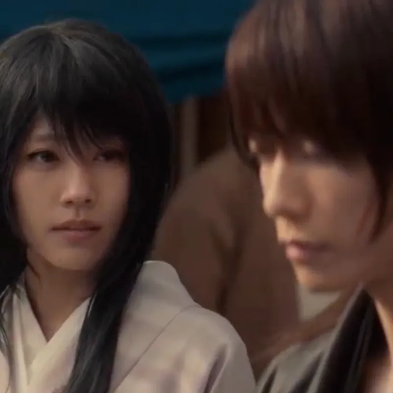 In this scene, Lady Tomoe is standing behind Kenshin, who's staring into a crowd of people. Lady Tomoe is wearing a light pink garment with a white lapel, and has a look of sadness in her. Kenshin is wearing a dark gray garment, and dispair is obvious on his face.
